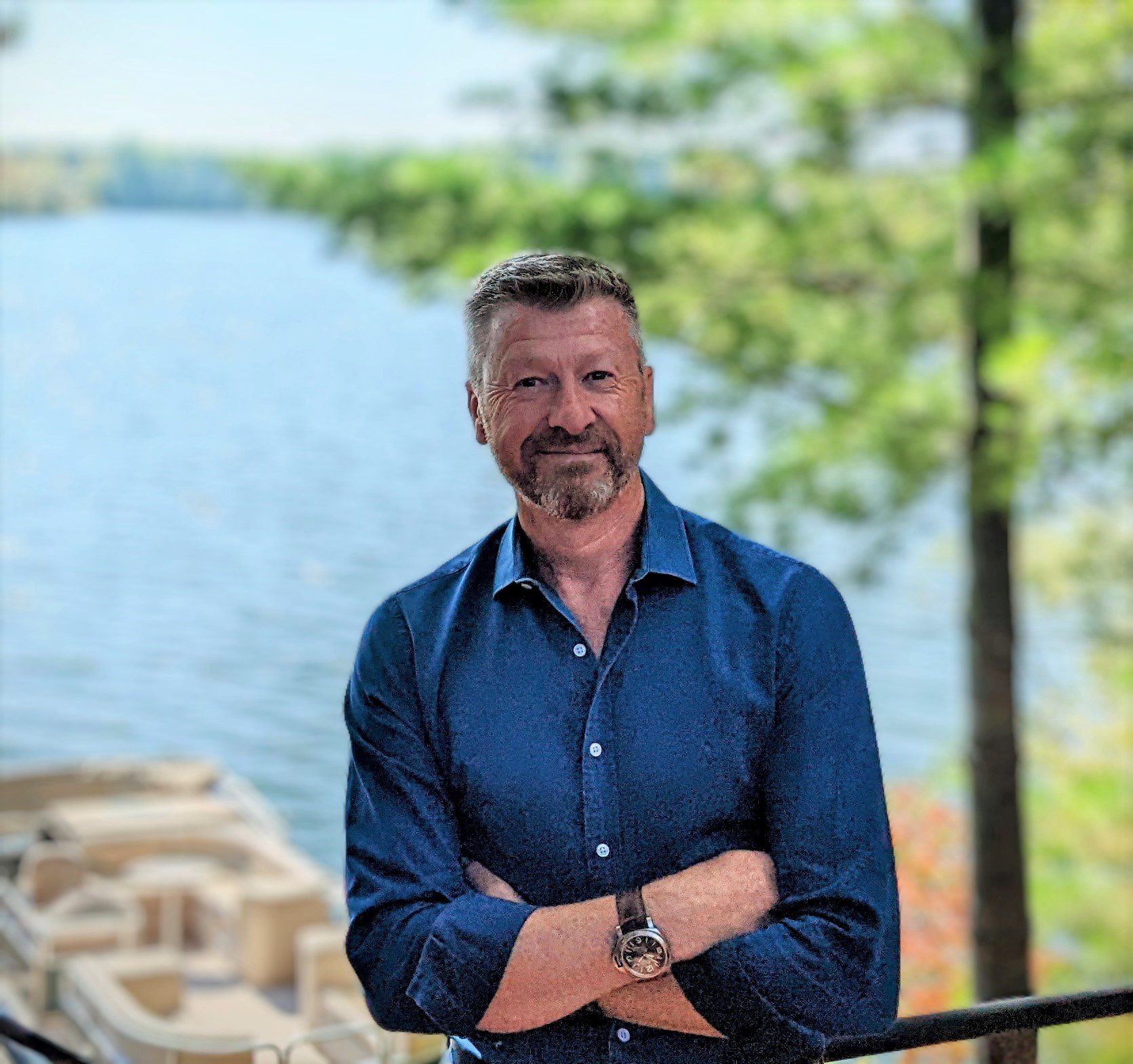Man with crossed arms stands by a lake, smiling.