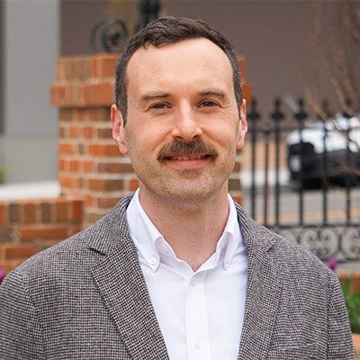 A man with a mustache wearing a gray blazer and white shirt outdoors.