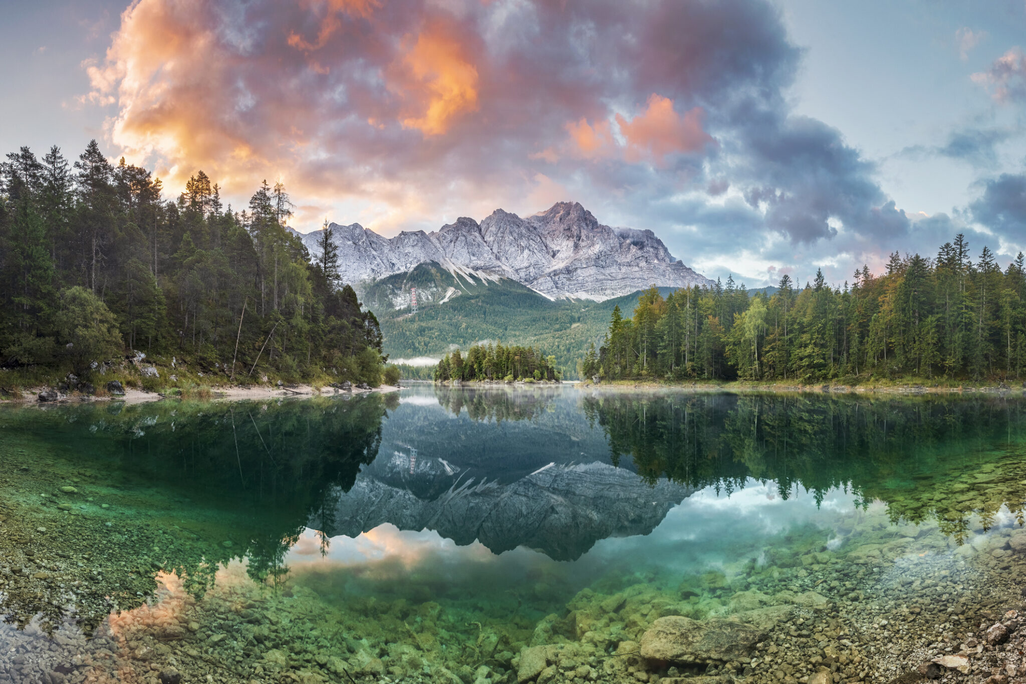 A serene lake reflects a colorful sunset sky and mountain range.