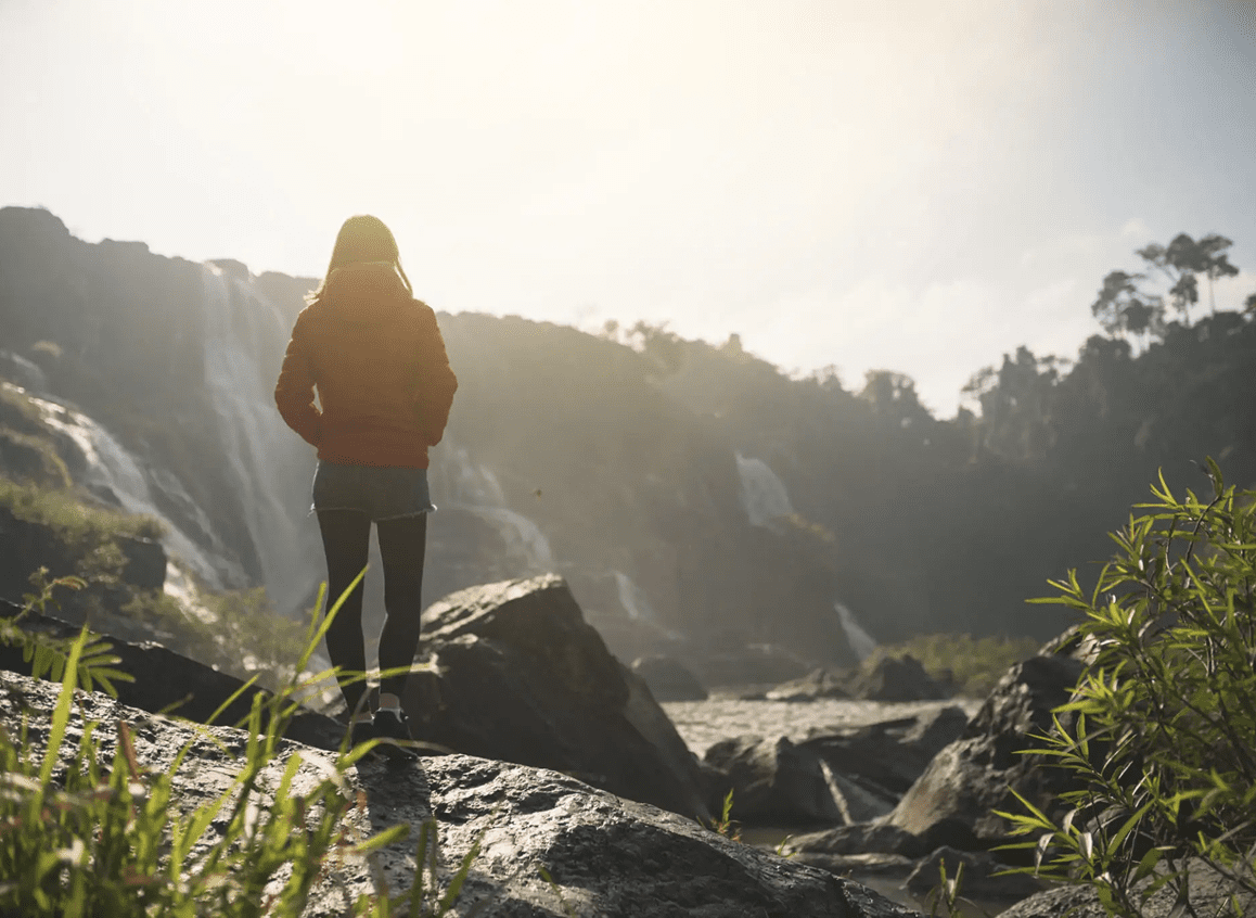 Person standing on rocks near a waterfall at sunrise.