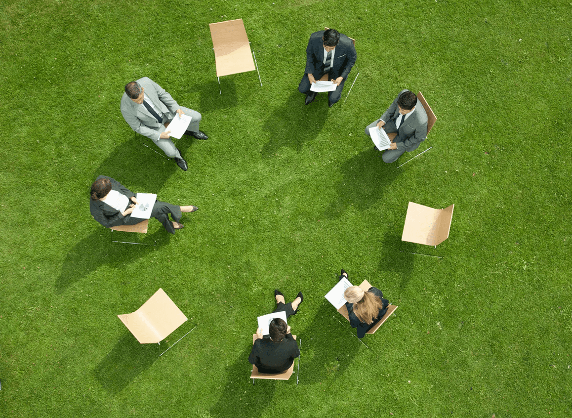 A group of six people sitting in a circle outdoors on grass, engaged in a discussion.