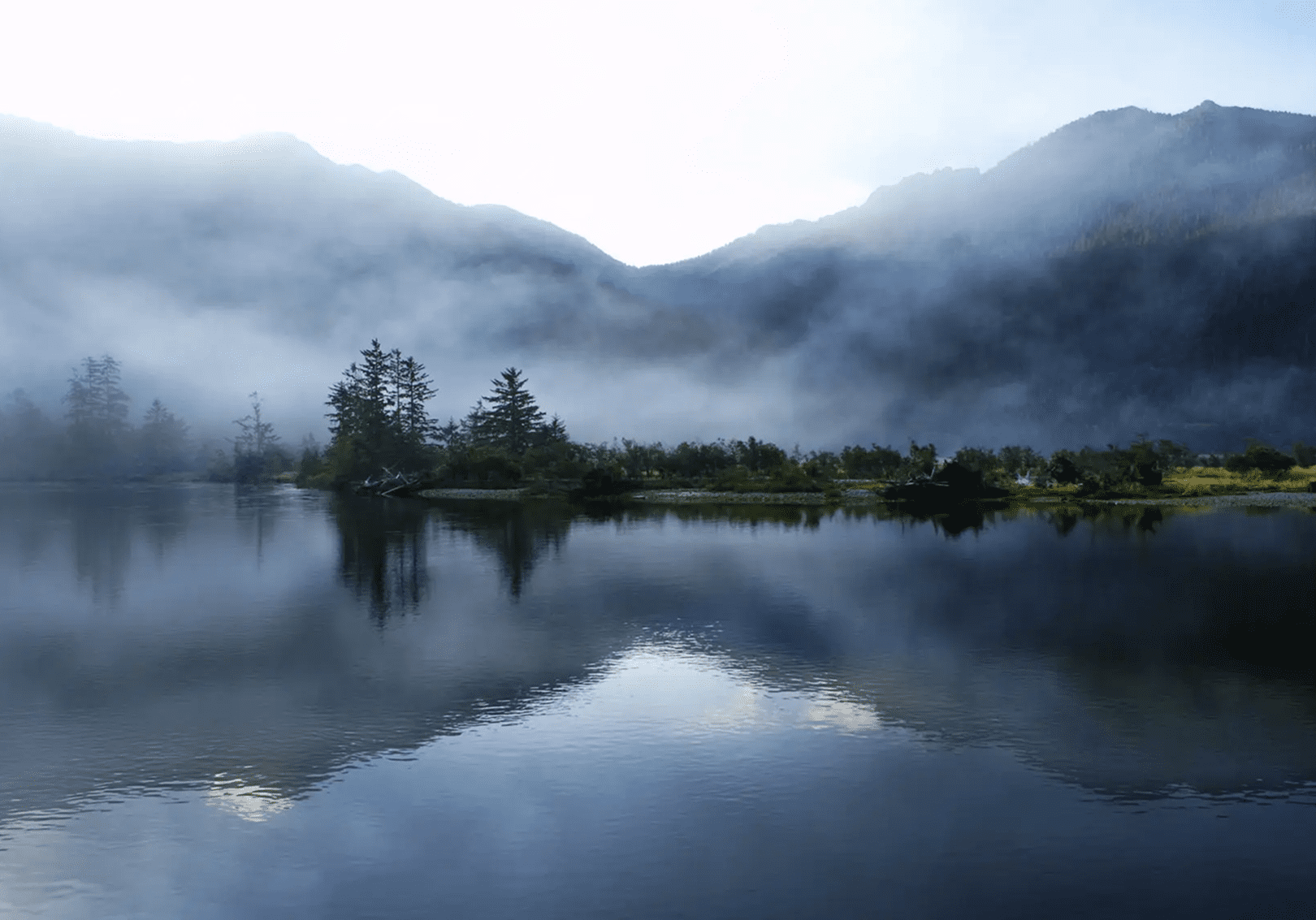 Mist-covered mountains reflected on a calm lake at dawn.