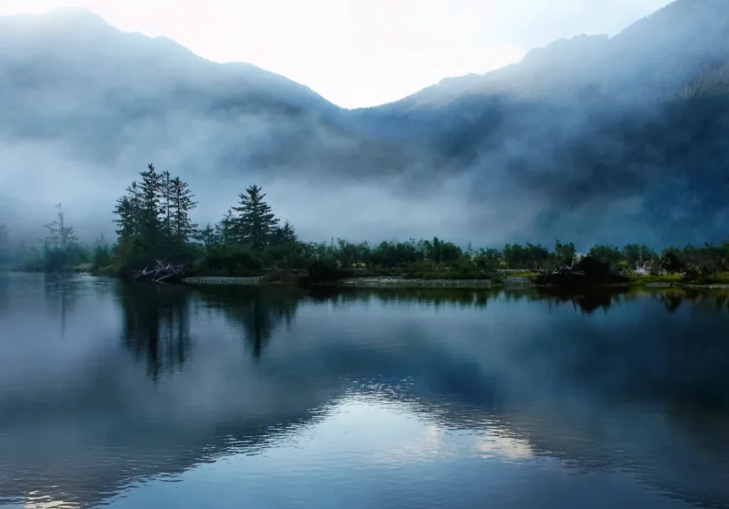 Mist-covered mountains reflected in a calm lake at dawn.