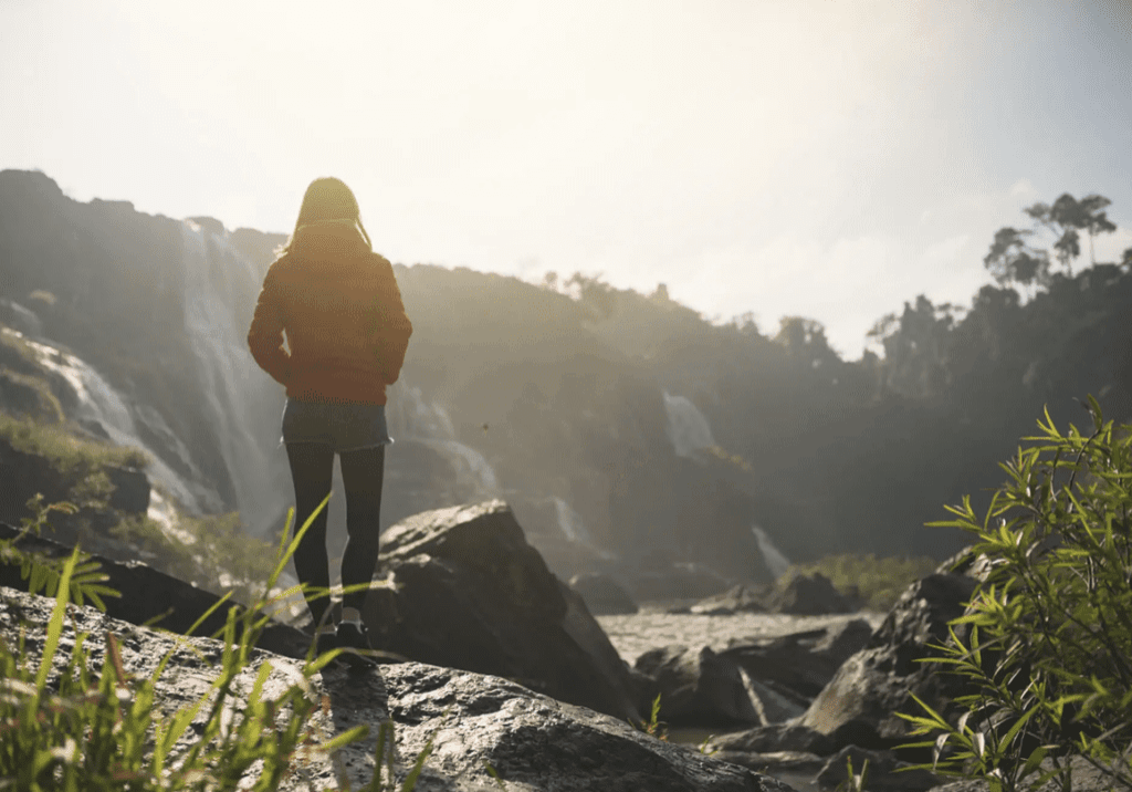 Person standing on rocks near a waterfall at sunrise.
