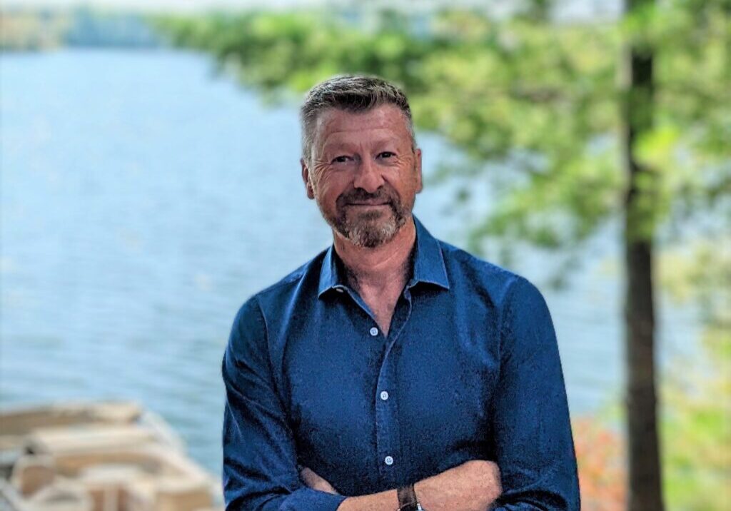 Man with crossed arms stands by a lake, smiling.