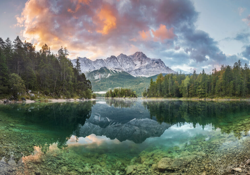 A serene lake reflects a colorful sunset sky and mountain range.