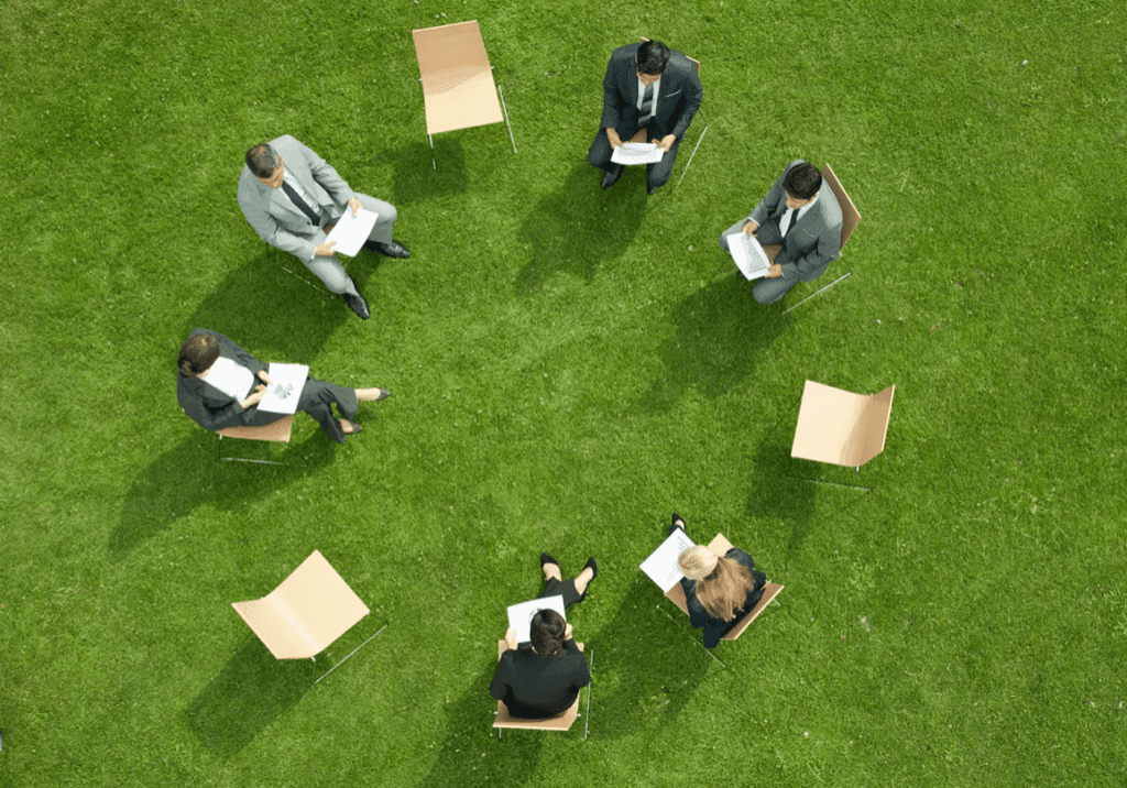 A group of six people sitting in a circle outdoors on grass, engaged in a discussion.